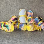 Seven packs of handmade festive mix makeup remover pads with colorful prints and yellow microfiber backing tied with yellow ribbons piled on grey felt