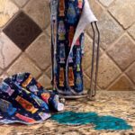 Muss Busters reusable towels in lava lamp cats print on paper towel holder with towel pulled off next to a spill on granite counter
