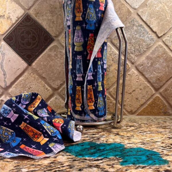 Muss Busters reusable towels in lava lamp cats print on paper towel holder with towel pulled off next to a spill on granite counter