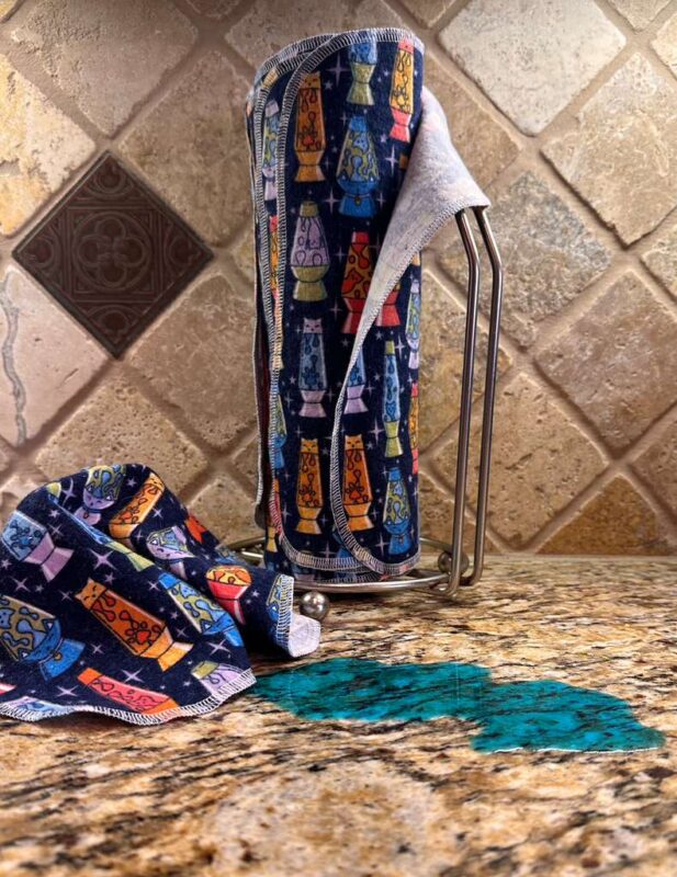 Muss Busters reusable towels in lava lamp cats print on paper towel holder with towel pulled off next to a spill on granite counter