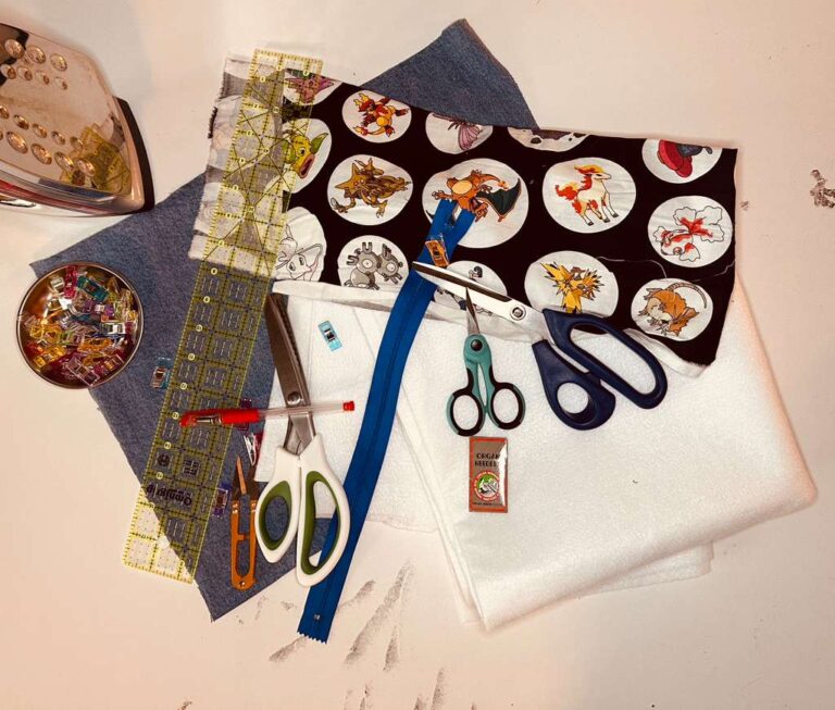 Flat lay of sewing supplies including denim, fabric, fusible fleece, zipper, scissors, clips, and tools on a cutting mat